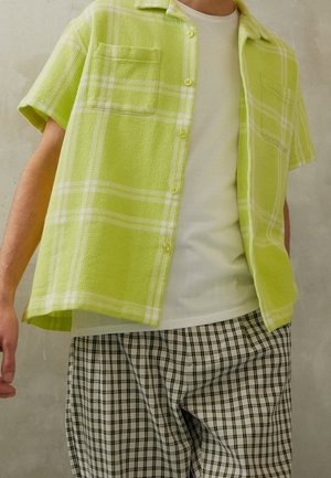 Personne portant une chemise à manches courtes à carreaux vert citron sur un t-shirt blanc avec un pantalon à carreaux noir et blanc sur un fond uni.