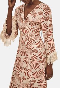 Vestido envolvente en crema con patrones abstractos en rojo y rosa. Presenta mangas anchas con acentos de flecos y una cintura ceñida. Tela de algodón.