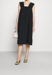 Černé bezrukávové šaty s volánovými ramenními akcenty, po kolena, v kombinaci se zelenými sandály na podpatku a světle růžovou kabelkou. Hladký textura.