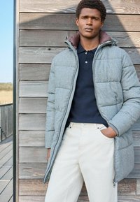 Chaqueta puffer gris con cuello marrón, presenta cierre frontal y diseño acolchado. Lleva puesta sobre una polo azul marino y combinada con pantalones crema.