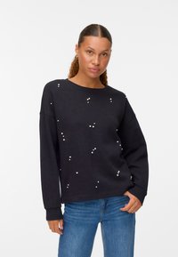 Sudadera negra de corte holgado, con detalles de perlas dispersas. Mangas largas y escote redondo, combinada con jeans azules.