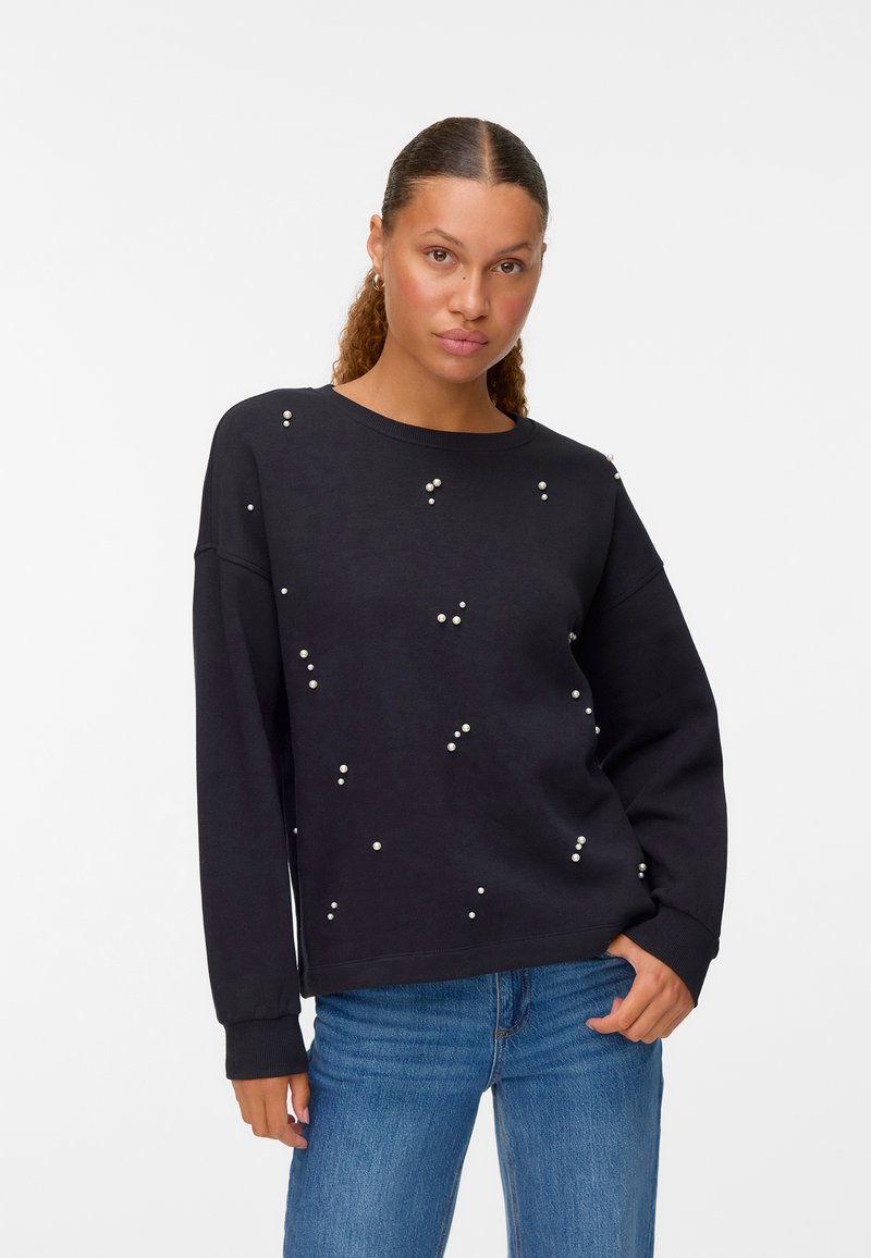 Sudadera negra de corte holgado, con detalles de perlas dispersas. Mangas largas y escote redondo, combinada con jeans azules.