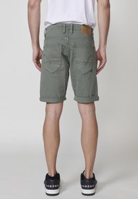 Shorts en denim vert olive avec des revers, poches arrière avec un design pointu, et une étiquette en cuir beige à la taille. Baskets noires.