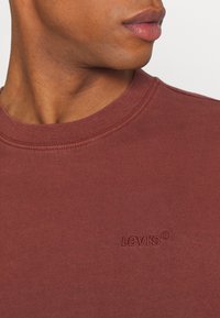 Sweatshirt à col ras du cou de couleur bordeaux, en coton. Présente un col côtelé et un logo "LEVI'S®" en relief sur la poitrine. Texture lisse, coupe décontractée.