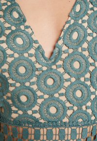 Teal broderade mönster med cirkulära motiv på en beige tyg. Har en V-ringning och intrikata spetsdetaljer på fåll.