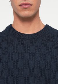 Námornícky modrý pletený sveter so štruktúrovaným vzorom, ktorý obsahuje vyvýšené štvorce, rebrovaný výstrih a hladký povrch.