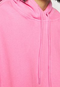 Rosa hoodie med dragsko, mjuk textur och avslappnad passform. Tyget verkar lätt och har synliga söm detaljer.
