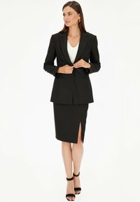 Czarna marynarka o dopasowanym kroju z klapami, jednobutonowa, oraz zestawiona z dopasowaną spódnicą do kolan z bocznym rozcięciem, uzupełniona czarnymi szpilkami.