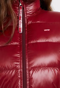 Rød pufferjakke med en blank overflade, høj krave og en lynlås foran. Har et lille Levi's-logo på venstre brystområde.