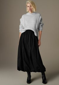 Maglione grigio lavorato a maglia con texture a coste, abbinato a una gonna maxi nera fluente. L'ambientazione presenta uno sfondo neutro e stivali alla caviglia.