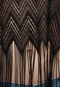 Μαύρο πλεκτό κορσές με ζιγκ ζαγκ μοτίβα και μακριά κρόσσια που αποκαλύπτουν γυμνή μέση πάνω από μπλε τζιν παντελόνια.