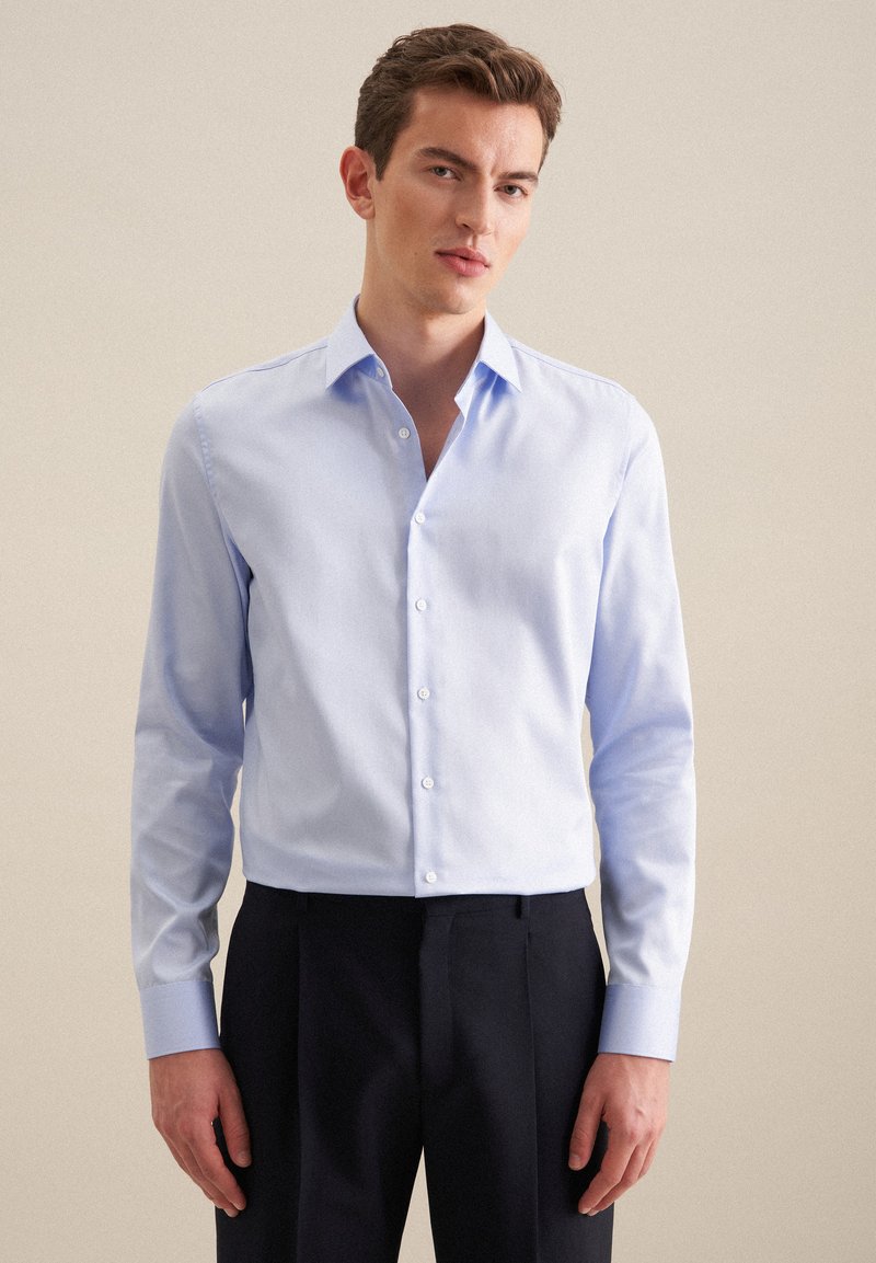 Jasnoniebieska koszula zapinana na guziki z wyraźnym kołnierzykiem, długimi rękawami i dopasowanym krojem. Gładka faktura i delikatny blask. Połączona z ciemnymi spodniami.