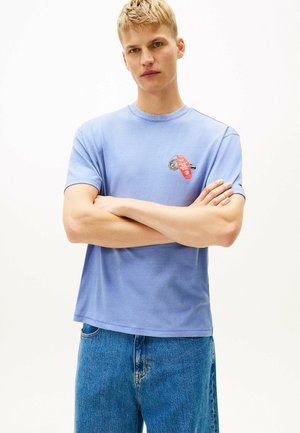 Helles blaues Baumwoll-T-Shirt mit kurzen Ärmeln, mit einem grafischen Druck eines roten und schwarzen Designs auf der Brust. Getragen mit blauen Jeans.