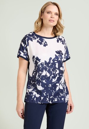 Blondynka w białej bluzce z krótkim rękawem ozdobionej granatowym kwiatowym wzorem oraz w granatowych spodniach, stojąca na tle jasnego jednolitego tła.