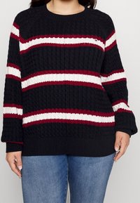 Musta kabelineulepaita, jossa on vaakasuoria viininpunaisia ja valkoisia raitoja, pyöreä kaula-aukko sekä resorit hihoissa ja helmassa. Paita on päälläsinisuurissa farkuissa.