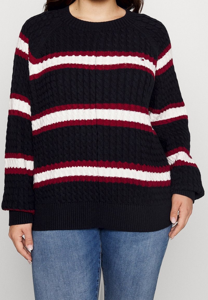 Czarny sweter w splot kablowy z poziomymi bordowymi i białymi paskami, okrągłym dekoltem oraz ściągaczami na rękawach i dole. Nosi się go na niebieskie dżinsy.