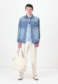 Light blue striped denim shirt with chest pocket, light blue t-shirt, cream trousers, and a cream tote bag. Sneakers with beige accents.