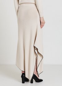 Jupe maxi beige tricotée avec une fente latérale et un bord festonné noir, en tissu doux, associée à des chaussures à talons noires.