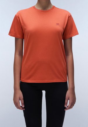 Osoba ubrana w jednolitą pomarańczową koszulkę z krótkim rękawem i czarne spodnie, stojąca na tle jednolitego jasnego tła.