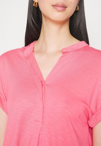 Rosa Kurzarmbluse mit Schlitzkragen, leichtem Stoff und vertikaler Nahtdetails. Glatte Textur mit lockerer Passform.