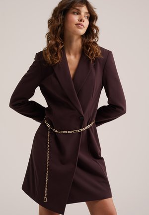 Femme aux cheveux ondulés portant une robe blazer brun foncé avec un bouton noir et une ceinture chaîne dorée, posant les mains dans le dos.