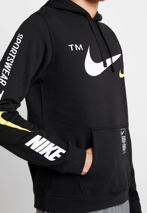 Person iført en sort Nike hoodie med hvide og gule logoer, "NIKE" tekst på ærmet og en lomme foran med et mærke.