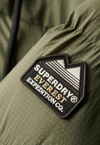 Tissu vert olive avec une texture matelassée, comportant un patch noir et blanc avec "SUPERDRY" et "EVEREST EXPEDITION CO." brodés.