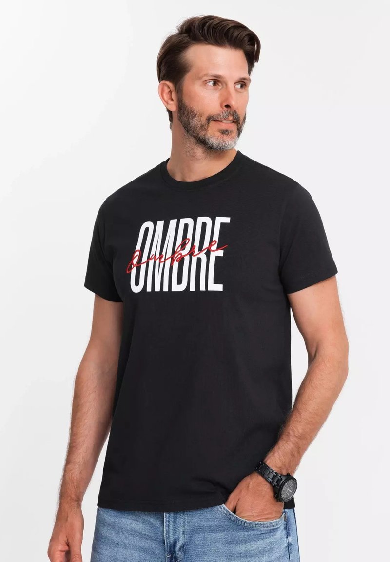 Camiseta de algodón negra con un gran texto blanco que dice "OMBRE" con un efecto de tachado en rojo. Diseño de mangas cortas y cuello redondo.