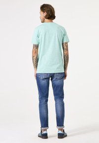 Camiseta de manga corta en verde menta, combinada con jeans azules arremangados y zapatillas con detalles azulados, exhibiendo tatuajes en los brazos.