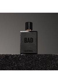 Bottiglia di profumo rettangolare nera texture con etichette "BAD" e "DIESEL" posata su una superficie nera ruvida su sfondo grigio scuro.