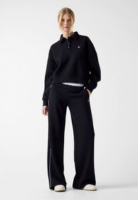 Black sweatshirt with a polo collar, cropped design, and emblem on the chest. Paired with wide-leg black pants featuring a side stripe.