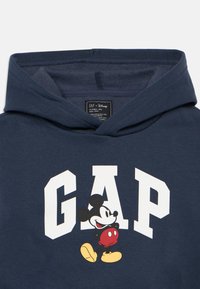 Tumši zila kapuce ar lieliem baltajiem burtiem "GAP", multfilmu pelītēm, kas tur sarkano bumbu, un mīkstu tekstūru. Iekļauta kapuce.