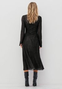 Schwarzes, langärmliges Kleid aus strukturiertem Stoff, mit einer fließenden Silhouette und geschwungenen Nahtdetails, kombiniert mit kniehohen schwarzen Stiefeln.