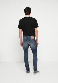 Čierne tričko s krátkym rukávom a tmavomodré úzke džínsy s hnedou nášivkou na zadnom vrecku, nosené so sivými teniskami.