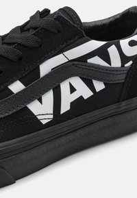 Gros plan sur une sneaker noire Vans avec un lettrage logo blanc en gras, des lacets noirs, une bande en cuir et un détail de semelle texturée.