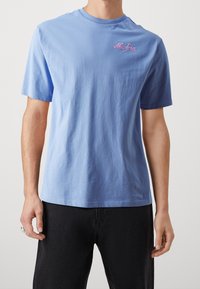 Světle modré bavlněné tričko s krátkými rukávy, kulatým výstřihem a vyšitým růžovým logem "New Era" na hrudi. Pohodlný střih a hladká textura.