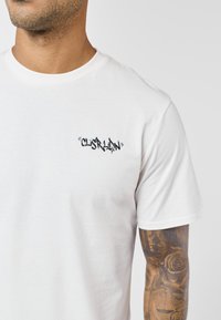 Mand iført almindelig hvid t-shirt med lille sort graffiti-lignende tekst på brystet og synligt tatoveringsærme med rose og vægte på højre arm.