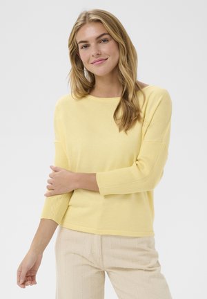 Jeune femme aux cheveux châtain clair portant un haut jaune à manches longues et un pantalon beige texturé, souriant avec un bras croisé sur son corps.