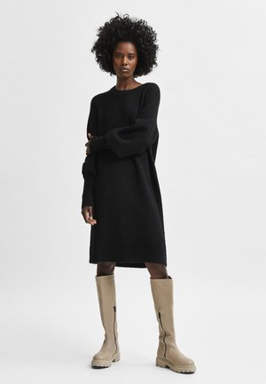 Schwarzes, gestricktes Oversize-Kleid mit langen Ärmeln, strukturierter Oberfläche und asymmetrischem Saum, kombiniert mit beigen kniehohen Stiefeln mit seitlichen Reißverschlüssen.