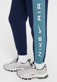 Námornícke tepláky so teal bočným pásom, na ktorom je biele logo "NIKE AIR", v kombinácii so striebornými teniskami Nike Air Max 97 a bielymi ponožkami.