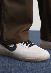 Sapatilha branca com swoosh preto, upper em camurça e couro texturizado, atacadores negros e sola de borracha transparente, combinada com calças castanhas.