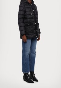 Chaqueta de plumas negra con capucha, botones de presión, dos bolsillos frontales y un acento texturizado negro. Combinada con jeans de pierna recta en lavado claro y botas negras de tobillo.