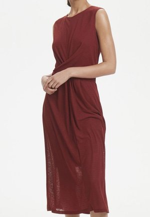 Mouwloze bordeaux midi-jurk met gedraaide voorkant, gedragen door een staande vrouw met de handen voor zich samengevouwen.