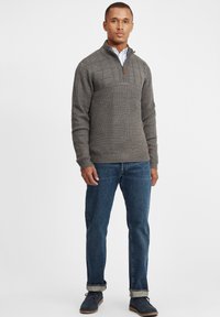 Grauer Strickpullover mit strukturiertem Muster und Viertelreißverschluss, kombiniert mit blauen Jeans und dunklen Schuhen, getragen von einem stehenden männlichen Modell.