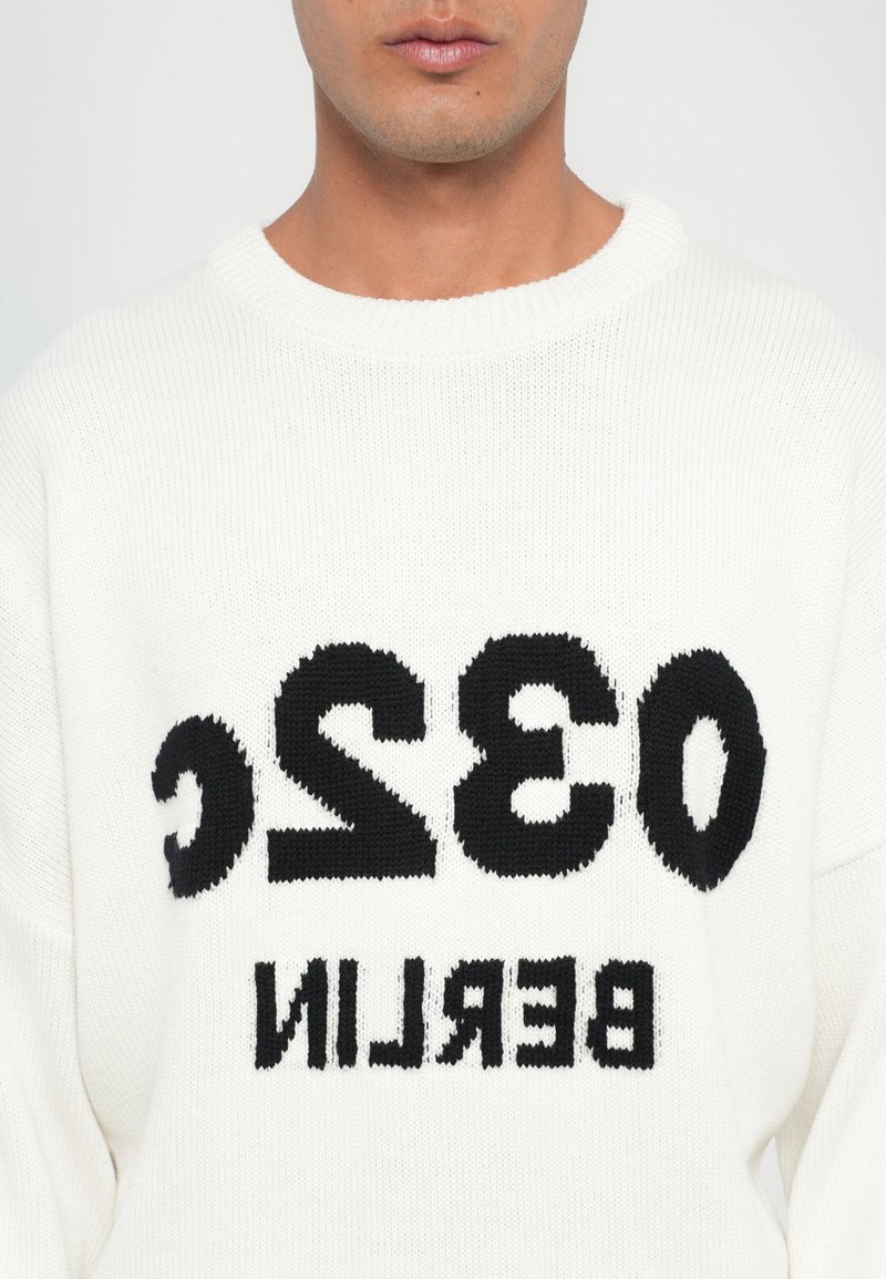 Pull en tricot blanc avec un grand texte noir écrivant "032 BERLIN" sur le devant. Le tissu a une apparence texturée avec une coupe ample.