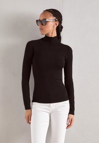 Black turtleneck sweater made of a fitted knit material, paired with white jeans. Model wears oversized sunglasses with a silver accent.