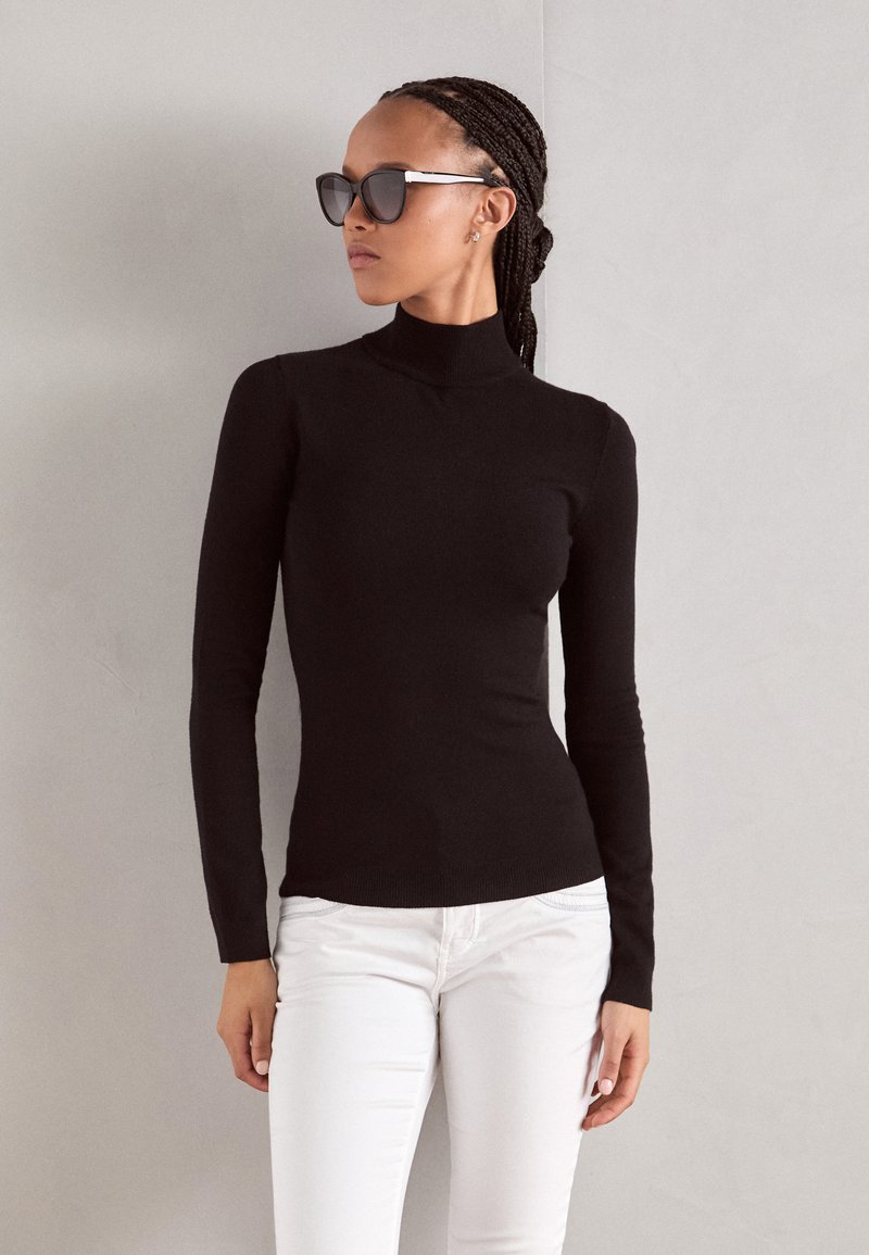 Black turtleneck sweater made of a fitted knit material, paired with white jeans. Model wears oversized sunglasses with a silver accent.