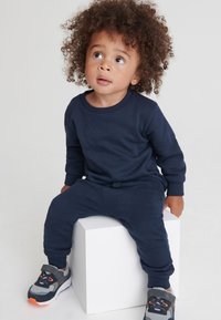 Maglietta a maniche lunghe in cotone blu navy con pantaloni abbinati, caratterizzata da polsini a coste. Completata con sneakers grigie e arancioni. Bambino seduto su un blocco bianco.
