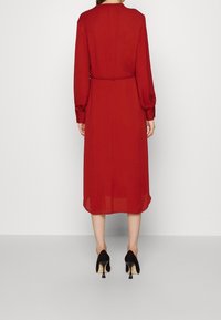 Rotes langärmliges Kleid mit einer taillierten Taille und einem fließenden Saum. Der Stoff wirkt leicht und leicht durchsichtig. Ein Paar schwarze High Heels.