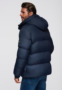 Granatowa kurtka puchowa z kapturem, charakteryzująca się pikowanym wzorem, ściągaczami przy mankietach oraz naszywką z logo na lewym rękawie. Dopasowany krój.